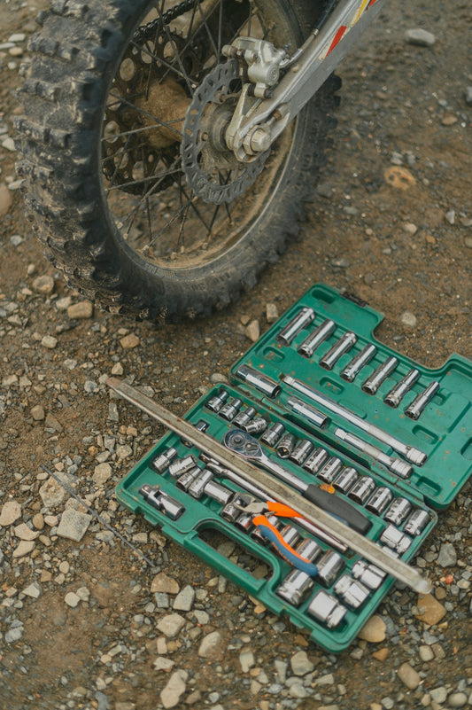 A green socket set and tools on rocky ground beside a motorcycle wheel